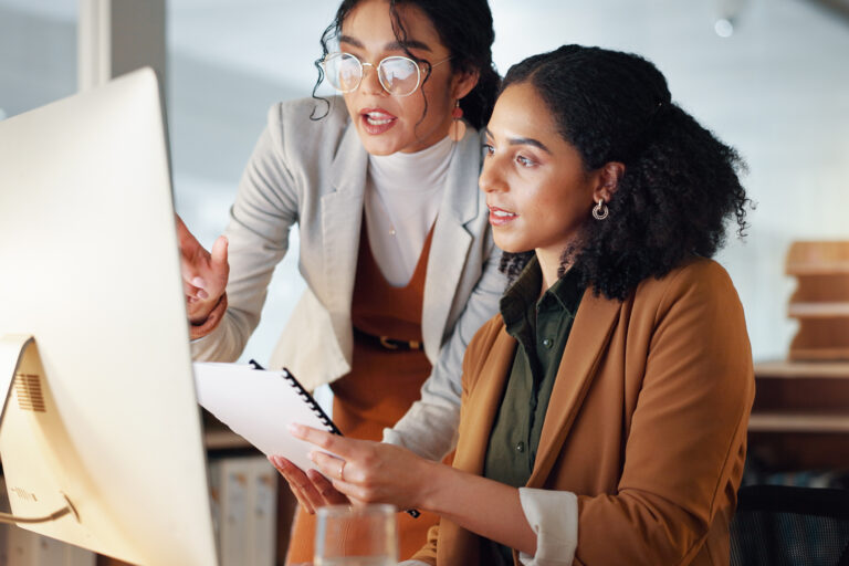 Two women collaborate at a computer, one gesturing while the other holds a notebook and looks at the screen.
