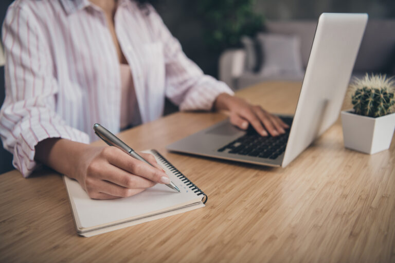 Person writing in a spiral notebook at a wooden desk, with a laptop and a small cactus plant nearby.
