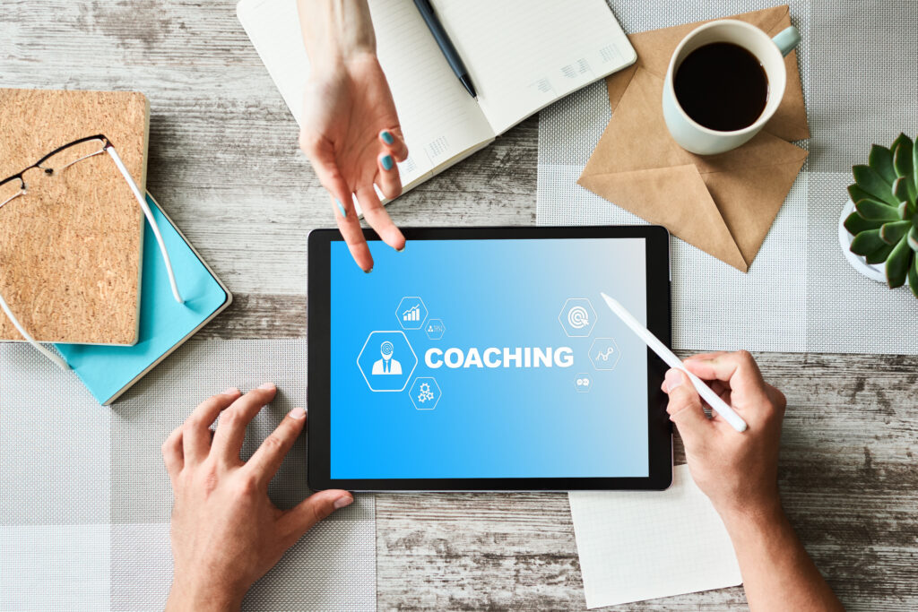 Tablet on a desk displaying a coaching app with hexagonal icons, surrounded by hands, a notebook, a coffee mug, and a plant nearby.